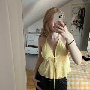 Gul ärmlös topp med knytband - Säljer en söt gul topp med tunna axelband och knytband framtill. Toppen är ärmlös och har en lätt volangdetalj vid nederkanten. Perfekt för varma sommardagar!