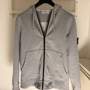 Grå hoodie från Stone Island - Säljer en stilren grå hoodie från Stone Island med dragkedja och justerbar huva. Tröjan har den ikoniska stone island loggan på ärmen och praktiska fickor framtill. Loggan på armen går att ta av och rekommenderas även att tas av vid tvättning. Den är knappast änvänd och är helt felfri. Hoodien är såklart äkta med ett Nypris på runt 4000kr mitt Pris är 1590kr. Hör gärna av er om ni är intresserade! 