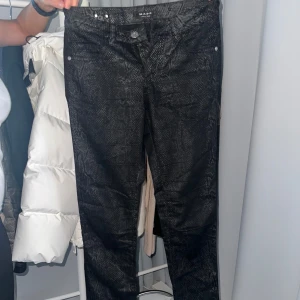 Svarta byxor med ormskinnsmönster från M.A.P.P - Snygga svarta byxor från M.A.P.P med ett coolt ormskinnsmönster. Byxorna har en knapp och dragkedja framtill samt fickor med silverfärgade detaljer. Perfekta för att ge din outfit en edgy touch. Nypris är 1395kr. De är i mycket bra skick. Tight passform, så snygga med exempelvis stövlar till. Model 3 och storlek 27