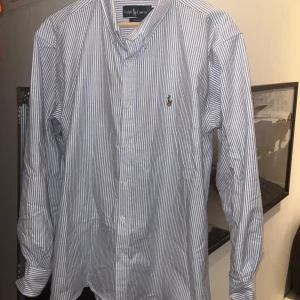 Randig skjorta från Ralph Lauren - Snygg och klassisk blå- och vitrandig skjorta från Ralph Lauren. Skjortan har långa ärmar och är broderad med den ikoniska logotypen på bröstet. Perfekt för en stilren look. Storleken är XL men passar även L men då lite baggy. Nypris 1749 kr. Kan mötas upp i Sthlm och pris kan diskuteras.