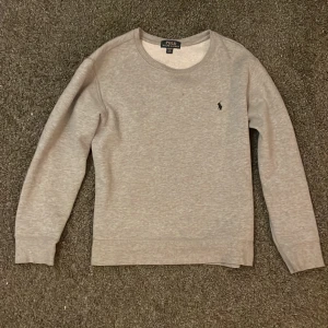 Grå tröja från Polo Ralph Lauren - Säljer en stilren grå tröja från Polo Ralph Lauren med rund hals och långärmad design. Tröjan har en liten broderad logotyp på bröstet. Den är helt i nyskick utan defekter. Köpt för 1100, säljes för 199. Storlek M barn (12y)