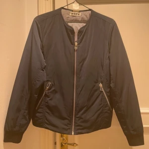 Blå bomberjacka från Hope - Snygg blå bomberjacka från Hope med dragkedja framtill och två snedställda fickor med dragkedjor. Jackan har en rund halsringning och långa ärmar. Perfekt för en stilren look.