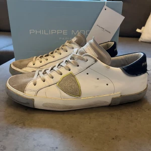 Vita sneakers från Philippe Model - Snygga vita sneakers från Philippe Model med grå detaljer och snörning. Skorna har en unik design med en kontrasterande svart häl och en grå tå. Perfekta för en stilren look. Sönder vid hälarna. Pris kan diskuteras.