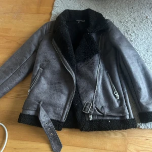 Grå skinnjacka med foder - Snygg grå skinnjacka med svart foder och dragkedjor. Jackan har en cool bikerstil med bälte i midjan och långa ärmar. Perfekt för att hålla värmen och se stilren ut samtidigt. Passar bra till både jeans och klänning. Nypris 899kr