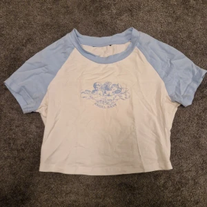 Vit och blå kortärmad topp med tryck - Säljer en söt vit och blå kortärmad topp med ett tryck av en ängel och texten 'Angel Baby' på framsidan. Toppen har en avslappnad passform och är perfekt för en casual look.