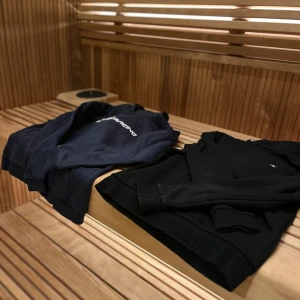 Svart och marinblå hoodie från Sail Racing - Två hoodies från Sail Racing, en i svart och en i marinblå. Båda har långa ärmar och en klassisk hoodie-design med justerbar huva. Perfekta för en avslappnad stil.