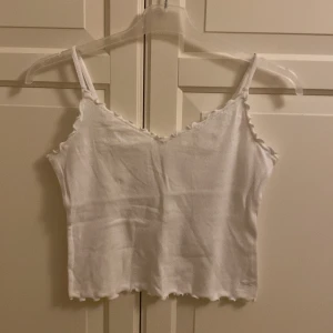 Vit ribbad linnetopp från Hollister - Säljer en söt vit ribbad linnetopp från Hollister. Toppen har smala axelband och en vågig kantdetalj vid halsringningen och nederkanten. I fint skick. Kort modell. Storlek L, men passar M. 