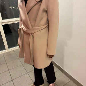 Beige kappa med bälte - Säljer en stilren beige kappa med bälte. Den har en elegant omlott-design och långa ärmar. Perfekt för kyligare dagar när du vill hålla stilen. Passar bra till både jeans och klänning.