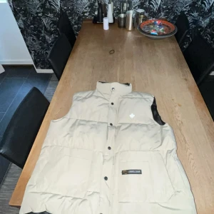 Beige dunväst från Canada Goose - Säljer en snygg beige dunväst från Canada Goose med broderad lönnlövdetalj på bröstet. Västen har en hög krage och knappar framtill. Perfekt för kyliga dagar när du vill hålla värmen utan att kompromissa med stilen. Västen är oanvänd och i nytt skick. Passar den som är 175 och längre