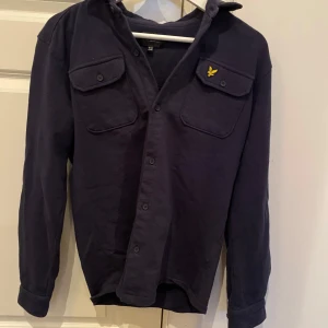Mörkblå skjorta från Lyle & Scott - Snygg mörkblå skjorta från Lyle & Scott med två bröstfickor och en gul broderad logga på ena fickan. Skjortan har långa ärmar och knappar framtill. Perfekt för en stilren look!