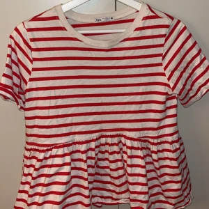 Randig topp från Zara - Säljer en söt randig topp från Zara i rött och vitt. Toppen har korta ärmar och en volangdetalj vid midjan som ger en fin form. Perfekt för en avslappnad stil.