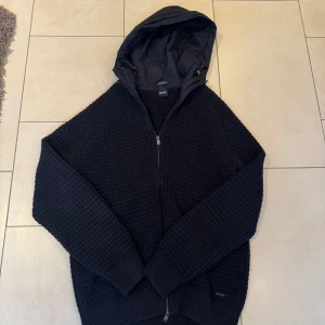 Mörk blå cardigan från Hugo boss - Säljer en stilren mörk blå  stickad cardigan från Boss med dragkedja och huva. Perfekt för kyliga dagar med sin mysiga och varma design. Cardiganen har långa ärmar och en ribbad nederkant för extra komfort.