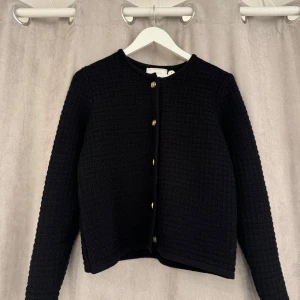 Neo Noir kofta - Snygg svart kavaj med guldknappar framtill. Den har en elegant och stilren design med långärmade ärmar och en rund halsringning. Perfekt för att ge en klassisk touch till din outfit.