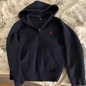 Mörkblå hoodie från Ralph Lauren - Säljer en snygg mörkblå hoodie från Ralph Lauren med dragkedja och röd logga på bröstet. Den har en klassisk passform med långa ärmar och en praktisk huva. OBS att den passar folk som är 13-14 år gammal också. Använd 1 gång.