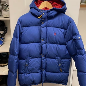 Blå dunjacka från Polo Ralph Lauren - Säljer en snygg blå dunjacka från Polo Ralph Lauren. Jackan har en huva och stängs med både dragkedja och knappar. Det är storleken L i barn storlekar och passar i 12-15 års åldern 