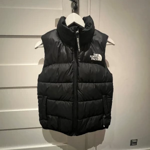 Svart dunväst från The North Face - Säljer en snygg svart dunväst från The North Face. Västen har en dragkedja framtill och en hög krage för extra värme. Logotypen är tryckt på bröstet och ryggen. Använt få gånger.