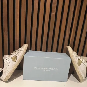 Vita sneakers från Philippe Mode - Snygga vita sneakers från Philippe Model med gulddetaljer och svart häl. Skorna har en klassisk design med snörning och en bekväm passform. Perfekta för en stilren look. Den har litet hål där bak.