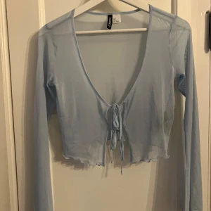 Ljusblå cardigan i mesh  - Säljer en ljusblå croppad topp från H&M. Toppen har långa ärmar och en knytning framtill. Materialet är tunt och ger en luftig känsla. Perfekt för en stilren och avslappnad look. Aldrig använd!
