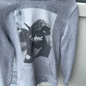 Grå hoodie med Tupac-tryck - Säljer en grå hoodie med ett stort svartvitt Tupac-tryck på framsidan. Tröjan har en klassisk passform med långa ärmar och en praktisk magficka. Perfekt för dig som älskar streetwear och vill ha en bekväm och stilren look.