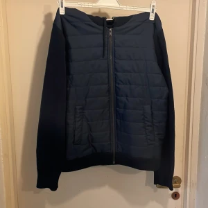 Mörkblå jacka med huva från Kappahl - Säljer en mörkblå jacka från Kappahl med huva och dragkedja. Jackan har quiltade detaljer på framsidan, stickat material i ryggen samt ribbade muddar vid ärmslut och nederkant. Perfekt för kyligare dagar.