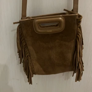 Brun mockaväska med fransar från Maje - Säljer en snygg brun axelväska i mocka från Maje. Väskan har coola fransdetaljer på sidorna och ett praktiskt handtag upptill. Perfekt för att ge din outfit en bohemisk touch.