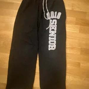 Svarta mjukisbyxor med tryck - Säljer ett par svarta mjukisbyxor med trycket '2018 Senior' i vitt på ena benet. Byxorna har en dragsko i midjan för justerbar passform. Perfekta för en avslappnad stil.