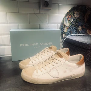 Beige, vita sneakers från Philippe Model - Snygga beige sneakers från Philippe Model med vita och bruna detaljer. Skorna har en klassisk design med snörning och en bekväm passform. Perfekta för en stilren vardagslook👌Använda under en sommar med omsorg till finare tillfällen🎩Orginal kartong och ettiket finns kvar! Hör gärna av dig vid frågor eller funderingar!