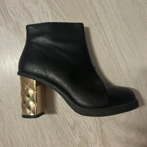 Svarta boots med gulddetaljer - Snygga svarta boots i skinn med en unik guldfärgad klack som har ett quiltat mönster. Skorna har en dragkedja på sidan för enkel på- och avtagning. Perfekta för att ge en elegant touch till din outfit.