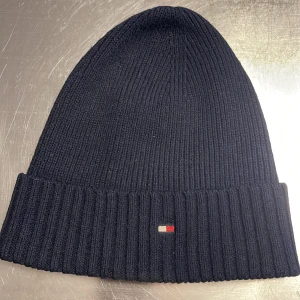Svart ribbad mössa från Tommy Hilfiger - Snygg svart ribbad mössa från Tommy Hilfiger med en liten logotypdetalj framtill. Perfekt för att hålla värmen under kyliga dagar.