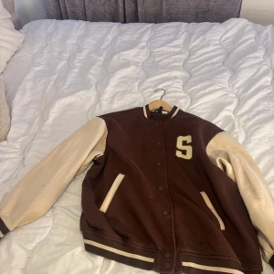 Brun och beige varsity jacka  med bokstav - Säljer en snygg brun och beige varsity jacka med bokstaven 'S' på bröstet. Jackan har långa ärmar och stängs med knappar framtill. Perfekt för en cool och avslappnad stil. Köpt för 600