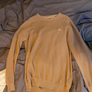 Beige stickad tröja från GANT - Säljer en stilren beige stickad tröja från GANT. Tröjan har en rund halsringning och långa ärmar, perfekt för kyligare dagar. Den är enkel att matcha med olika outfits och ger en klassisk look.
