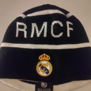 Mössa - Snygg svart och vit mössa med RMCF-text och Real Madrid-logga framtill. Perfekt för att visa ditt stöd för laget under kalla dagar. Mössan har ett klassiskt randigt mönster och är tillverkad i ett mjukt material för extra komfort.