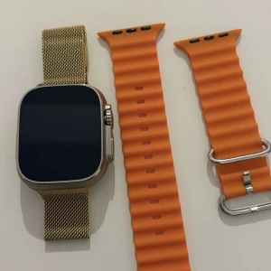 Appel watch 2  - Snygg smartklocka med en fyrkantig skärm och två utbytbara armband. Ett elegant guldmetallarmband och ett sportigt orange silikonarmband ingår. Perfekt för både stil och funktionalitet som har en egen laddare.