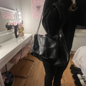 Zara väska  - Snygg svart axelväska i skinn med kedjedetaljer på axelremmen. Väskan har en praktisk dragkedja upptill för säker förvaring. Perfekt för att ge en stilren touch till din outfit.