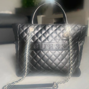 Guess väska i skinn - En svart Guess väska i skinn material med kedjedetaljer och dragkedja. Perfekt för att bära dina dagliga nödvändigheter med stil. Väskan har ett rymligt inre med flera fack för enkel organisering.