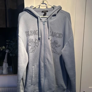 Ljusblå hoodie från Blinglucid - Säljer en ljusblå hoodie från Blinglucid med dragkedja och huva. Tröjan har ett tryck med texten 'BLINGLUCID' och två änglar på framsidan. Perfekt för en avslappnad stil.