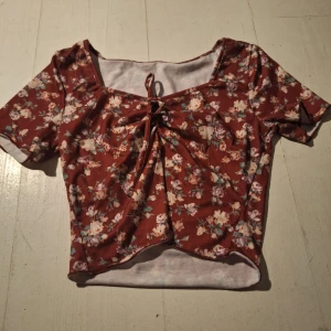 Blommig röd topp med knyt - Säljer en söt röd topp med blommigt mönster och knytning framtill. Toppen har korta ärmar och en fin fyrkantig halsringning. Perfekt för en somrig look!