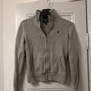 Grå stickad zip tröja från Polo Ralph Lauren - Säljer en stilren grå stickad tröja från Polo Ralph Lauren med dragkedja och broderad logotyp på bröstet. Tröjan har en hög krage med ribbade detaljer i rött och blått samt långa ärmar. Perfekt för kyligare dagar.skick 8/10 storlek XS