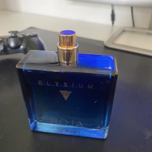 Elysium Parfym från Roja Parfums - Säljer en lyxig Elysium parfym från Roja Parfums. Flaskan är elegant i en djupblå färg med guldiga detaljer och en fyrkantig form. Perfekt för den som vill ha en exklusiv doftupplevelse.