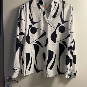 Vit och svart mönstrad skjorta - Snygg vit blus med abstrakt svart mönster. Blusen  har långa ärmar och en stilren design utan krage. Perfekt för en trendig look.