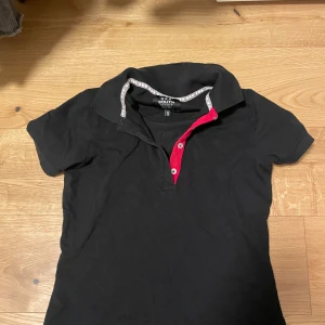 Svart pikétröja från South west - Snygg svart pikétröja från South west med korta ärmar och en kontrasterande röd knappslå. Perfekt för en avslappnad stil. Tröjan har en klassisk krage och är tillverkad i ett bekvämt material.