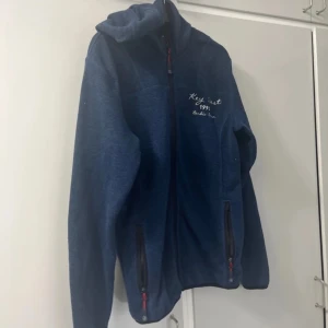 Blå fleecejacka från Key West Medium - Blå fleecejacka från Key West med broderad text på bröstet. Storlek Medium,  dragkedja och två fickor med dragkedjor. Nopprar finns.
