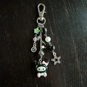 Nyckelring med Kuromi och stjärndetaljer - Charmig nyckelring med en Kuromi-figur, dekorerad med pärlor och en stjärna. Den har en mix av svarta, vita och genomskinliga pärlor samt en liten rosa rosett på Kuromi. Perfekt som accessoar för att ge din väska eller nycklar en personlig touch.