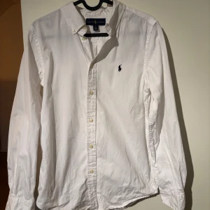 Vit skjorta från Ralph Lauren - Klassisk vit skjorta från Ralph Lauren med broderad logga på bröstet. Skjortan har långa ärmar och knappar framtill. Perfekt för en stilren look.