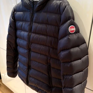 Svart pufferjacka från Colmar - Säljer en snygg svart pufferjacka från Colmar. Jackan har en dragkedja framtill och en huva för extra skydd mot väder och vind. Perfekt för kyliga dagar. Den har långa ärmar och en stilren design med Colmar-logga på ärmen. 