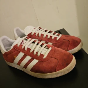 Röda Adidas Gazelle sneakers - Snygga röda Adidas Gazelle sneakers i mocka med vita detaljer och snörning. De har den klassiska tre-randiga designen på sidorna och Adidas-loggan på hälen. Perfekta för en stilren och sportig look. Lådan finns tyvärr inte kvar, billigare affär vid snabb affär 