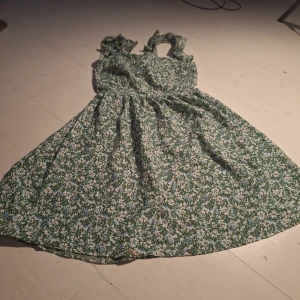 Grön blommig klänning - Charmig grön klänning med ett blommigt mönster i vitt och blått. Klänningen har volangdetaljer och en knytning framtill. Perfekt för sommarens varma dagar.