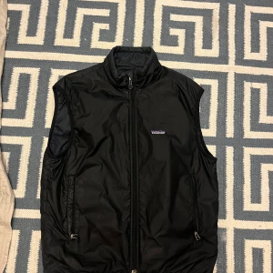 Svart väst från Patagonia - Säljer en stilren svart väst från Patagonia. Västen har en dragkedja framtill och är perfekt för lager-på-lager under kyligare dagar. Den är ärmlös och har en hög krage för extra värme. Passar bra till både vardag och utomhusaktiviteter.