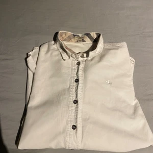 Vit dam skjorta från Burberry - Säljer nu min vita skjorta från Burberry är en elegant och stilren half button-modell för damer. Den är i storlek L, men jag märkte att den passar mer som en S eftersom jag råkade tvätta den i för hög temperatur, vilket gjorde att den krympte lite. Modellen på bilderna är 167 cm lång och brukar vanligtvis ha storlek S i skjortor, så det ger en bra referens för passformen. Skjortan är 100% äkta och är perfekt för både vardag och mer formella tillfällen. Materialet är 100% av burberrys fina bomull