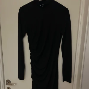 Svart långärmad klänning - Snygg svart långärmad klänning med en elegant drapering på sidan. Perfekt för en kväll ute eller en fest. Klänningen har en tight passform som framhäver figuren. Ett måste i garderoben för alla som älskar stilrena plagg!